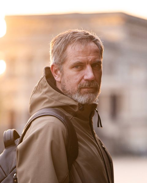 Un homme d’une cinquantaine d’années, barbe grise, sac à dos sur l’épaule, dans une rue au soleil couchant.